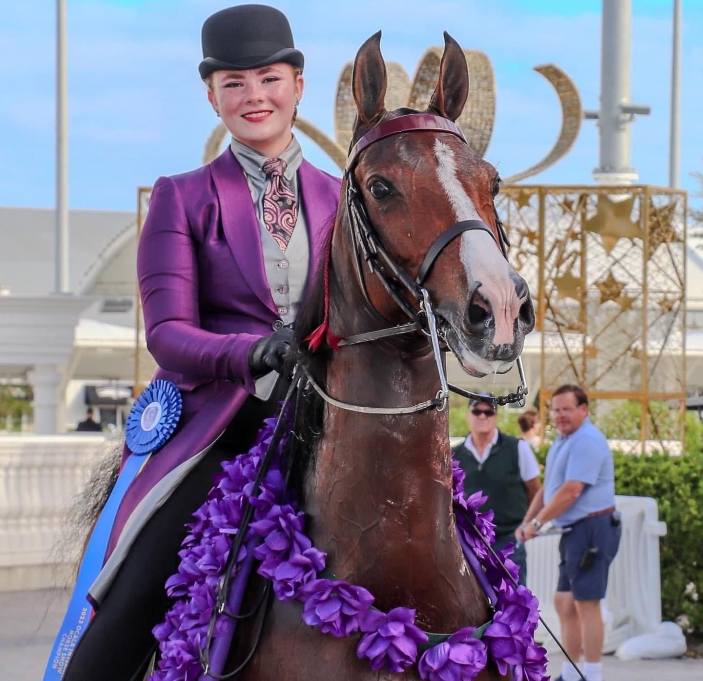 Photo of girl wearing a purple riding coat and blue ribbon sitting on an American Saddlebred horse with a garland of purple flowers draped over horse