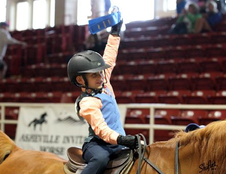 photo of young Lenux Stables rider holding blue ribbon in air while leaving arena at horse show