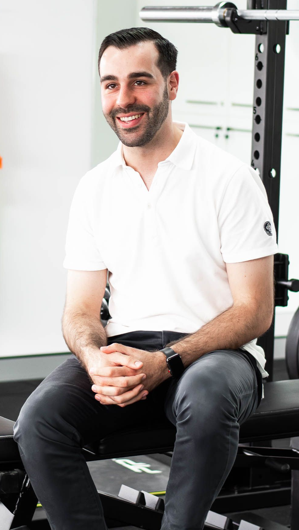 Man smiling, seated on bench in gym, wearing white polo shirt and gray pants.