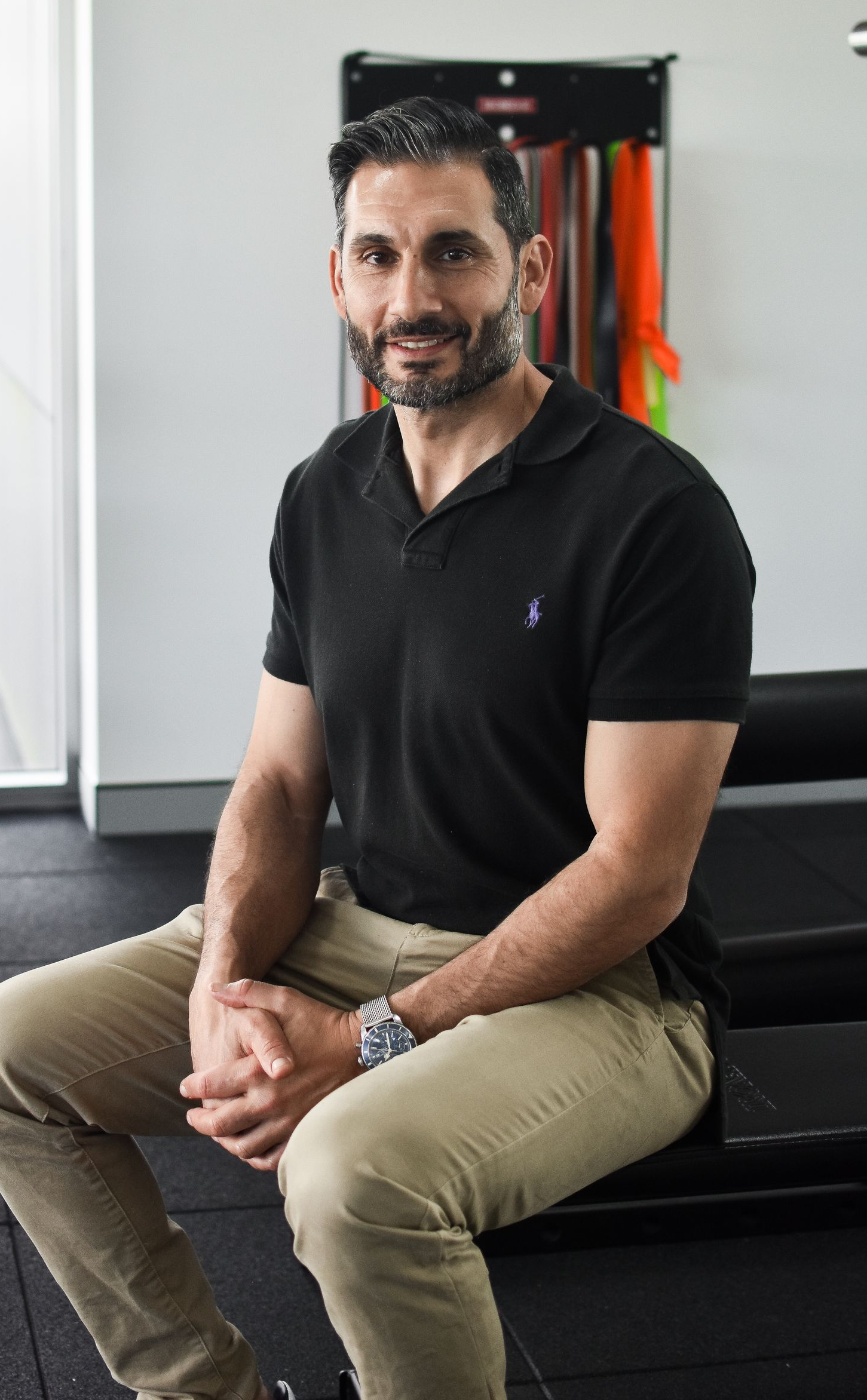 Man in black polo shirt and khaki pants, sitting on a gym bench, looking at the camera.