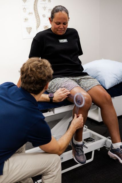 Physiotherapist measuring a patient's knee with a goniometer. Patient sits on a bed.