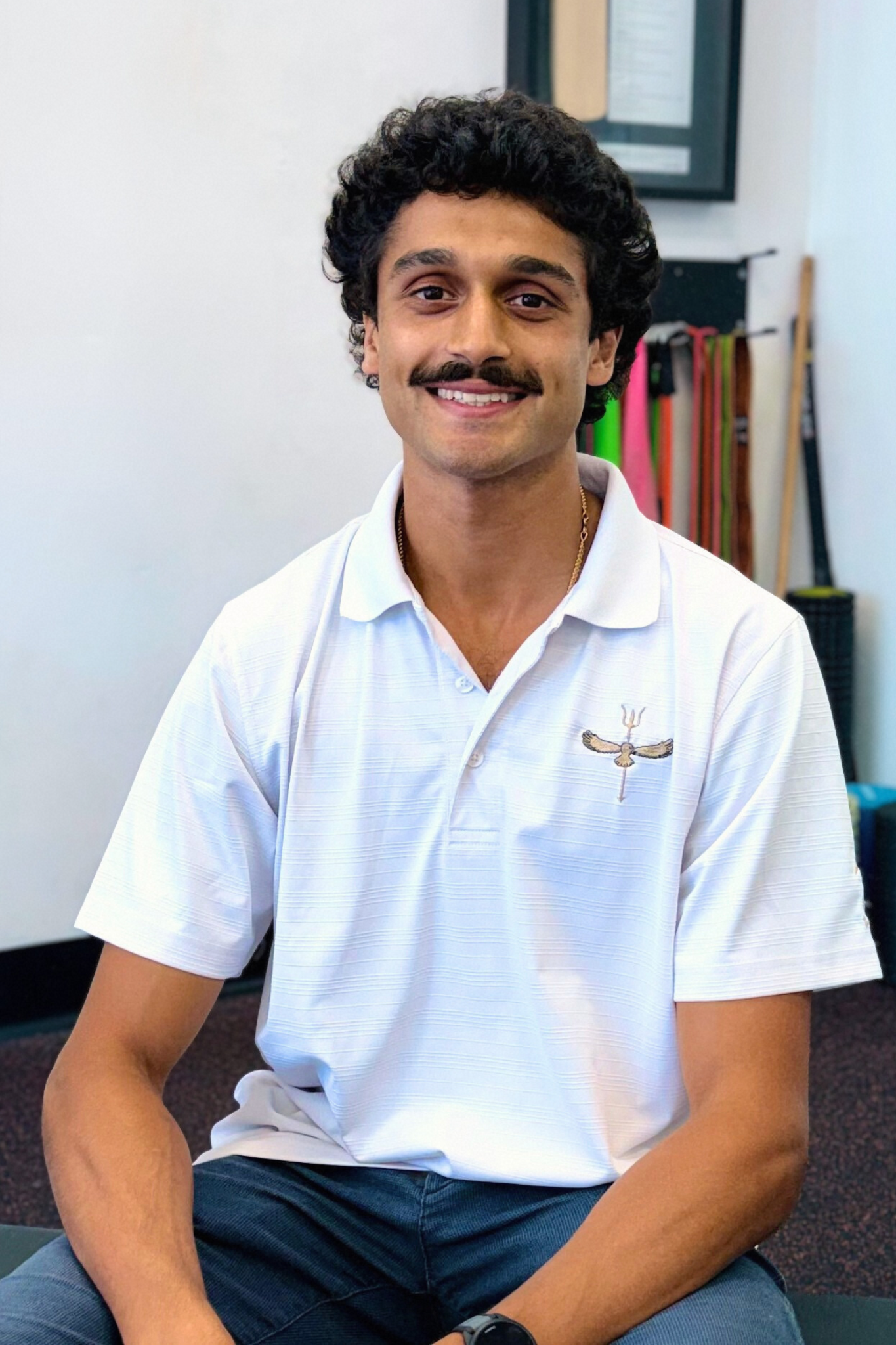 Man with dark curly hair and mustache smiles, wearing a white polo shirt, sitting indoors.