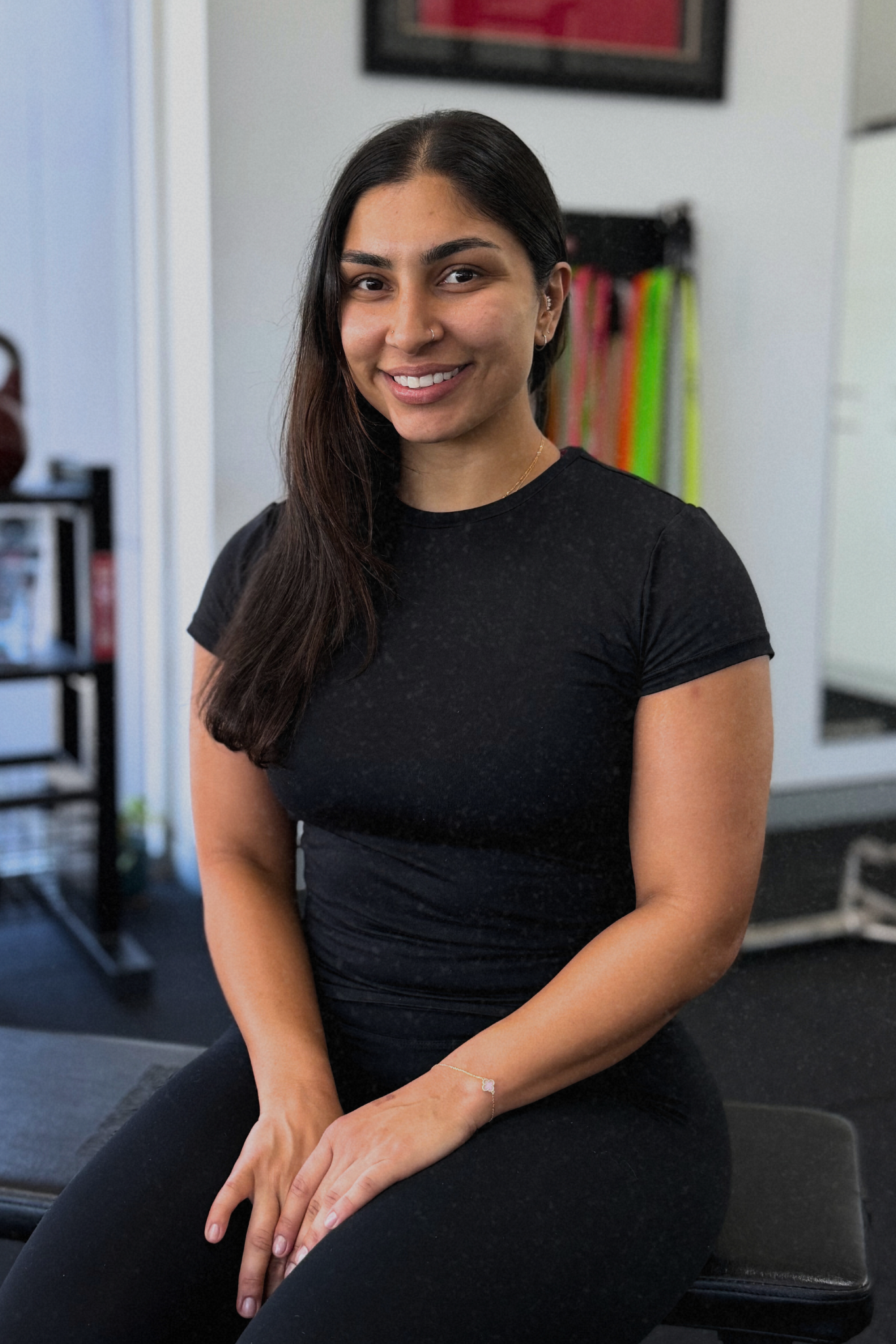 Woman with dark curly hair smiles, seated on a bench in a gym, wearing black clothing.