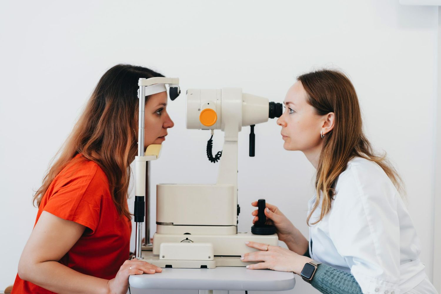 Two women facing each other across an eye exam machine in a white clinic room
