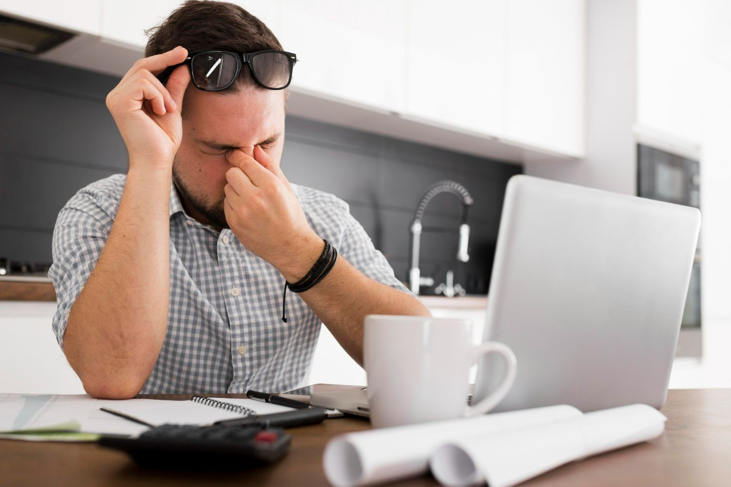 Man at desk with laptop, pinching his eyes, looking stressed. Coffee mug, papers, and calculator are on the table.