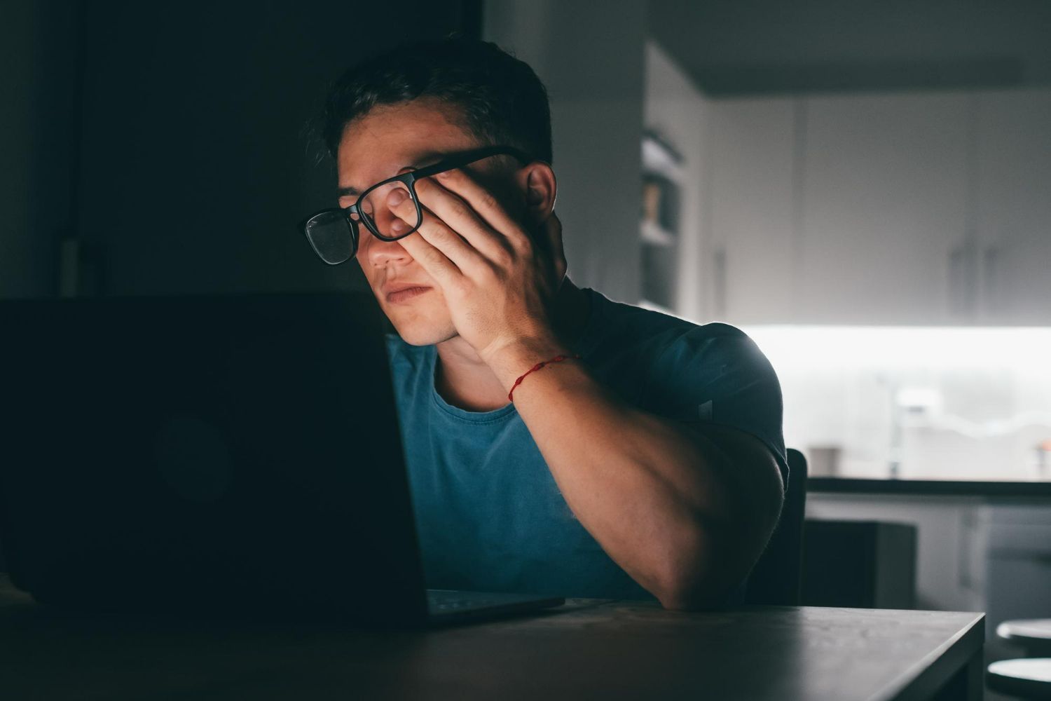 Person at a laptop in a dim kitchen, rubbing their eyes in a tired, late-night moment