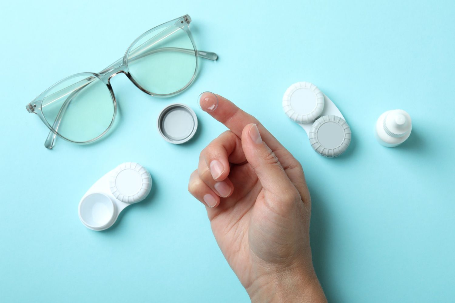 Hand with a drop of liquid, contact lens case, glasses, and eye drops on a blue background.