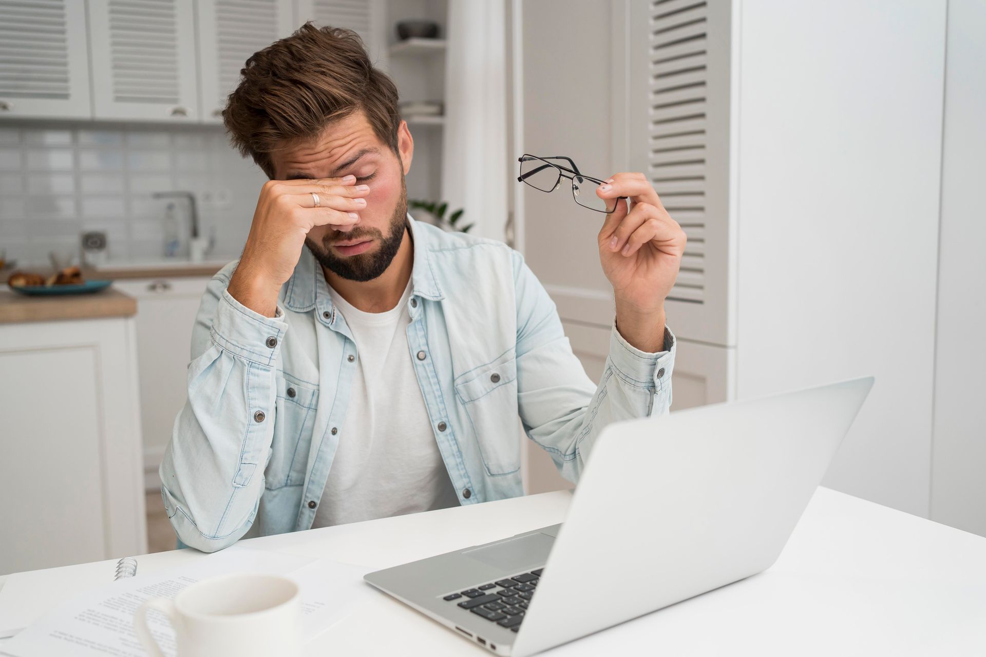 A man is sitting at a table with a laptop and rubbing his eyes.