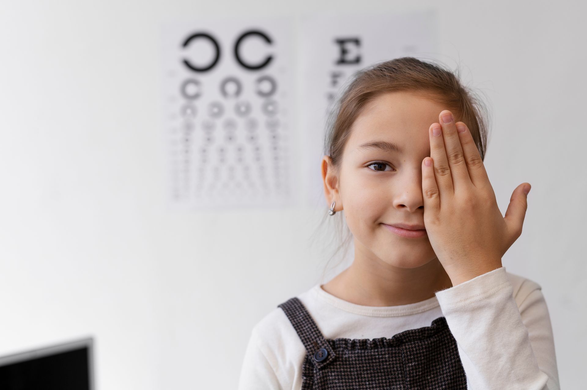 Girl with hand over one eye, in front of eye chart, smiling.