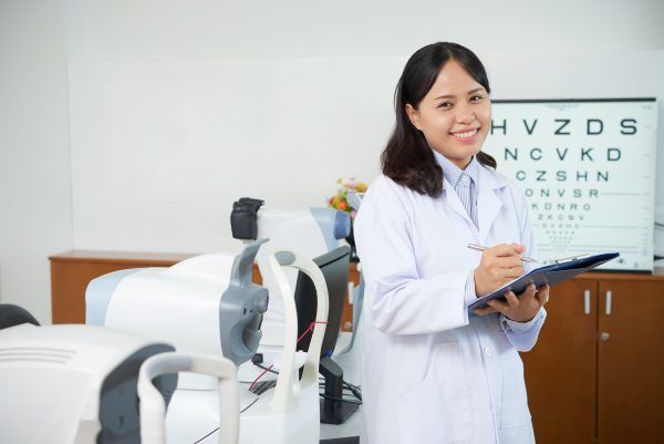 A female ophthalmologist is holding a clipboard and smiling in front of an eye chart.