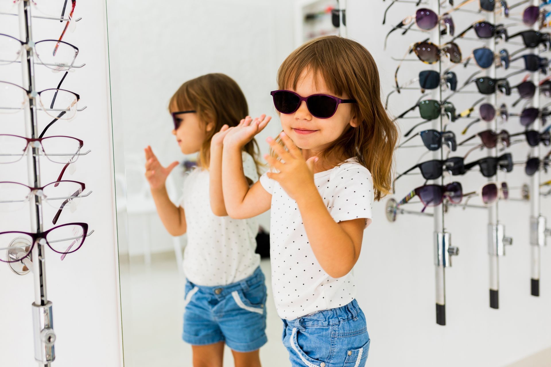 A little girl wearing sunglasses is standing in front of a mirror in an optical store.