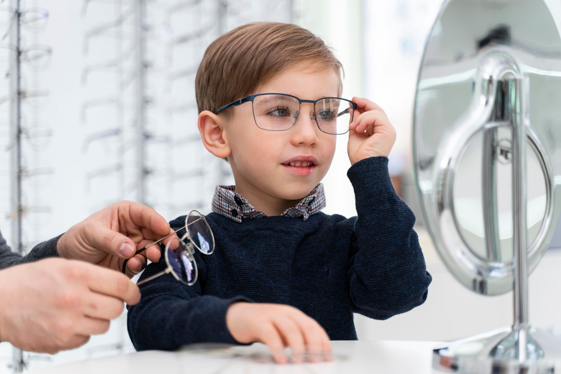 A young boy is trying on glasses in front of a mirror.