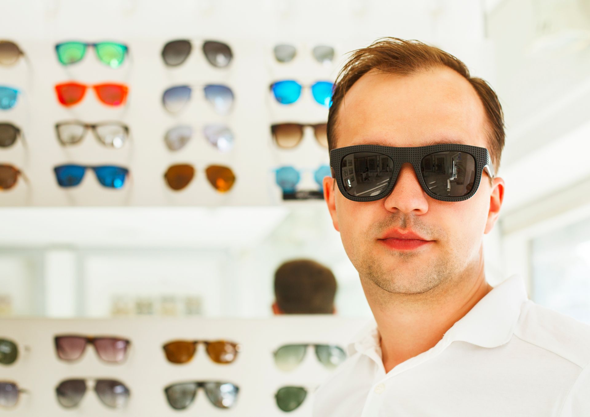 A man wearing large black sunglasses poses in front of a display of sunglasses in a shop.