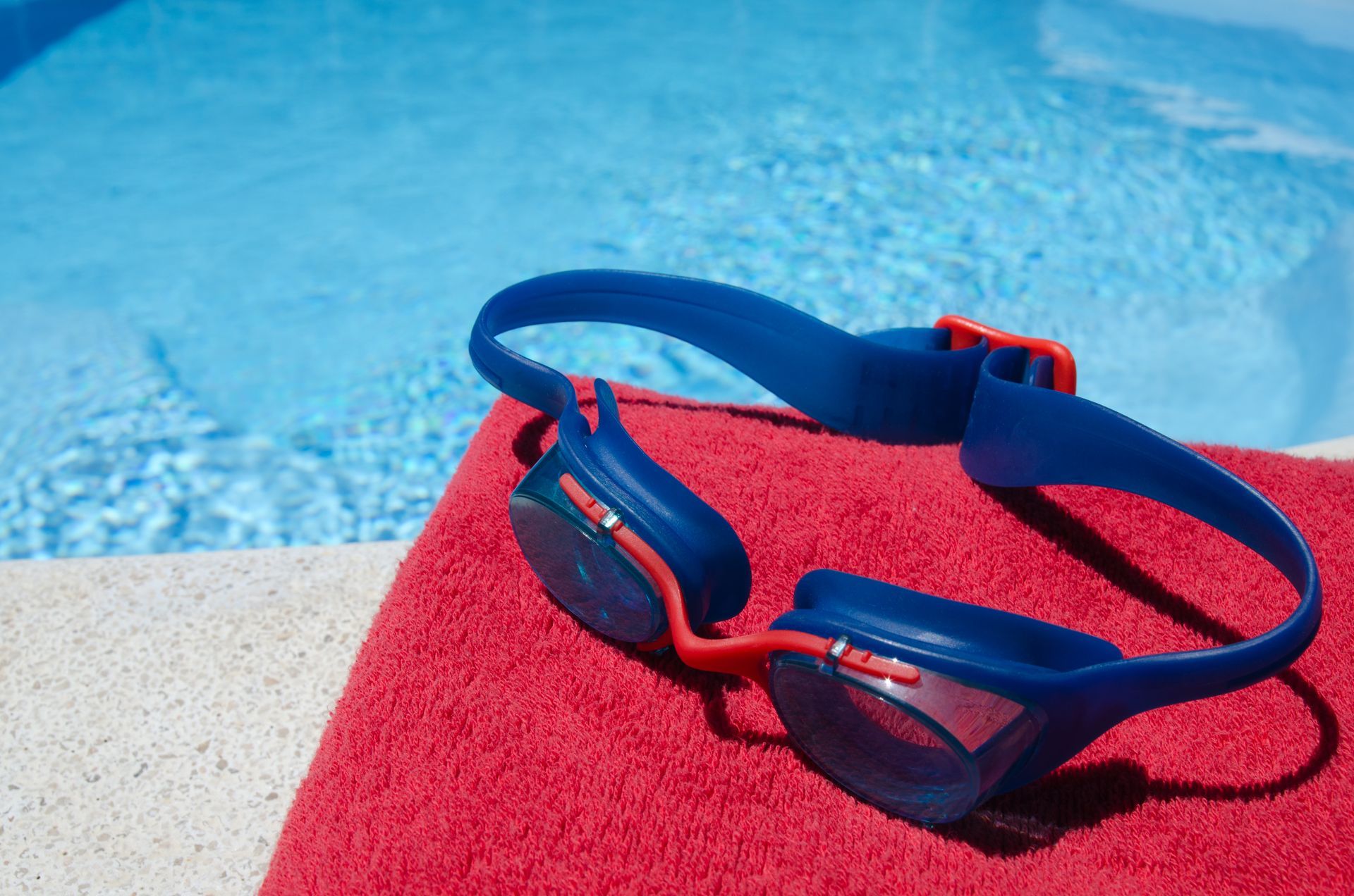 A pair of swimming goggles sitting on top of a red towel next to a swimming pool.