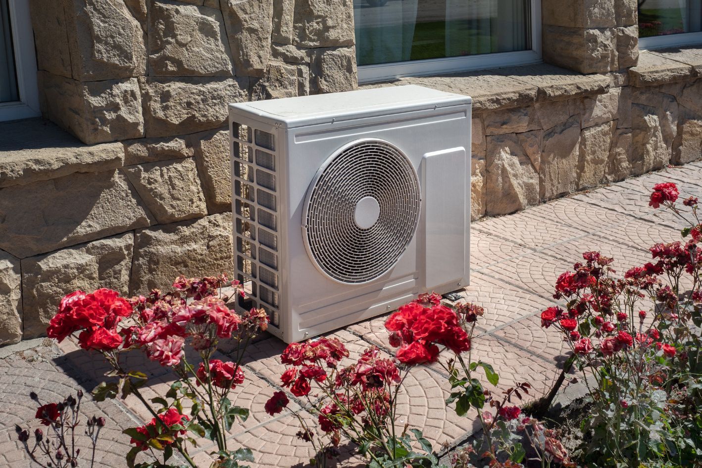 A small heat pump outside a beige stone house with some red flowers in front of it.