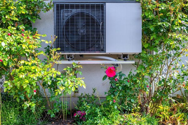 An air source heat pump with a grey body and black fan, installed around green plants on the wall of a property in Exeter, Devon.