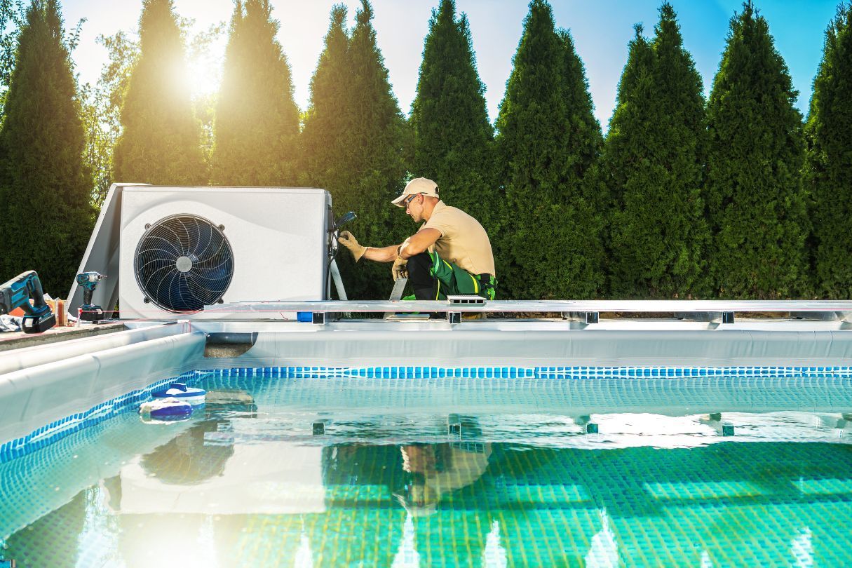 A man installing a white heat pump next to a swimming pool surrounded buy trees