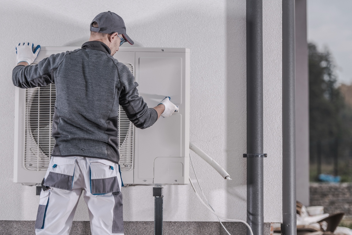 A man wearing a grey jumper and hat and white trousers installing a heat pump.