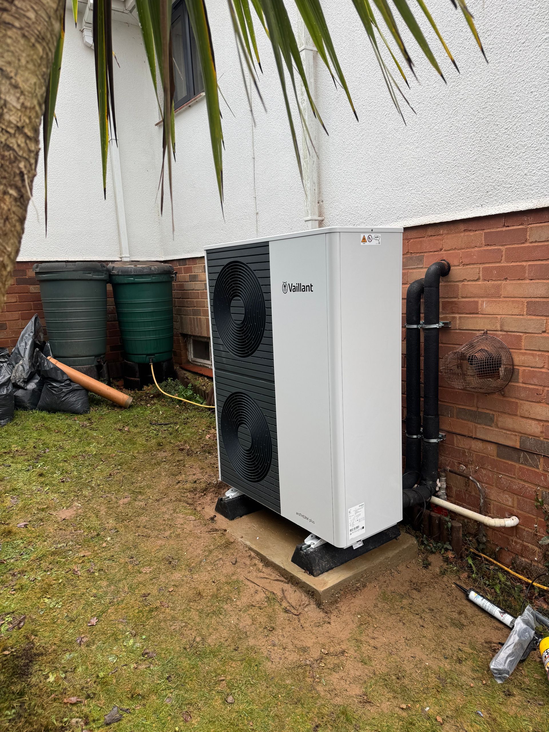 Outdoor heat pump unit beside a brick wall on a grassy yard, with pipes and a palm tree nearby.