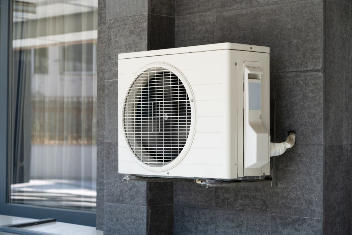 A white air conditioner is mounted on the side of a building next to a window.