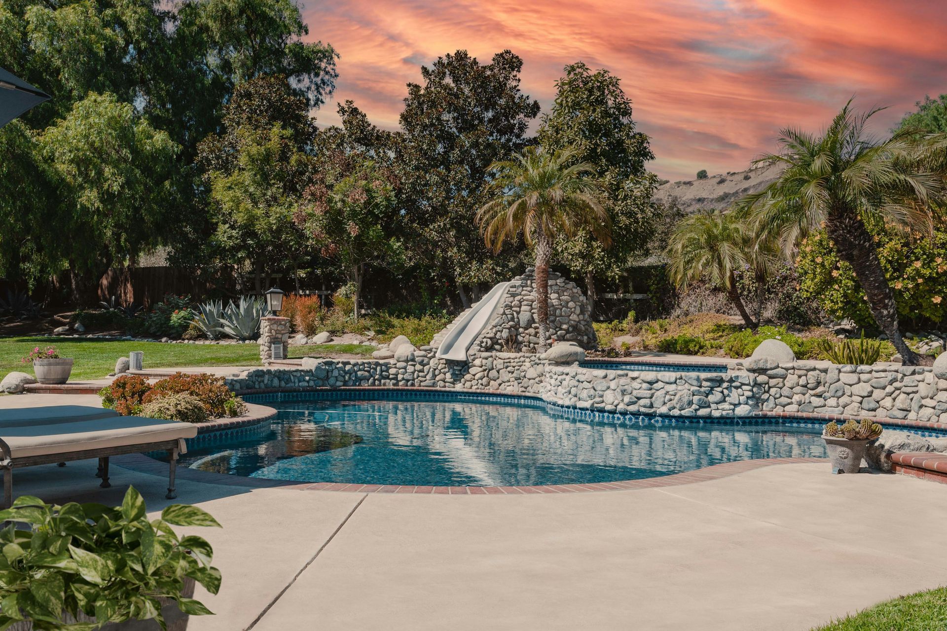 Backyard with pool, rock waterfall, palm trees, green lawn, and orange sunset sky.