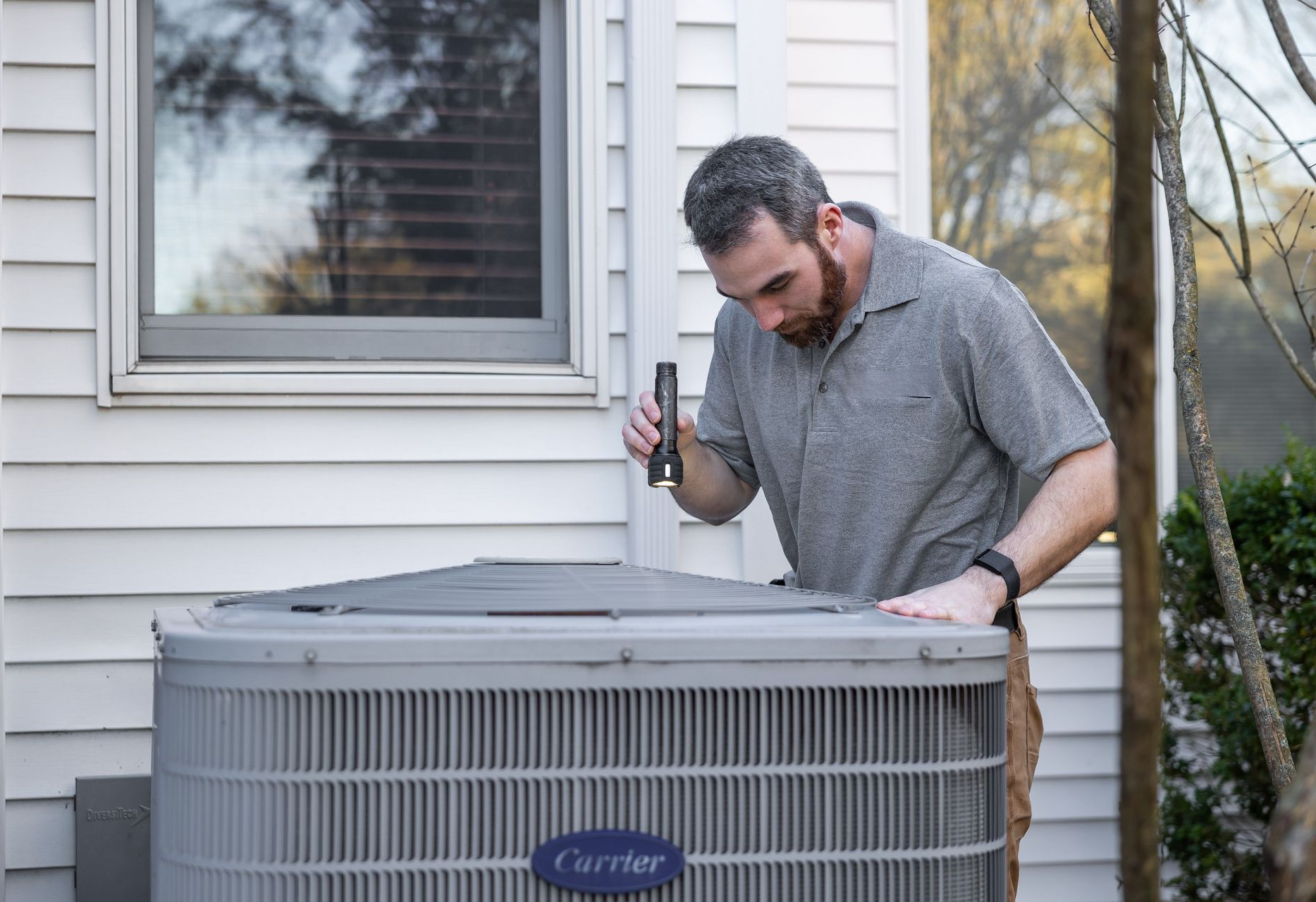 Man inspecting an outdoor air conditioner unit beside a house window