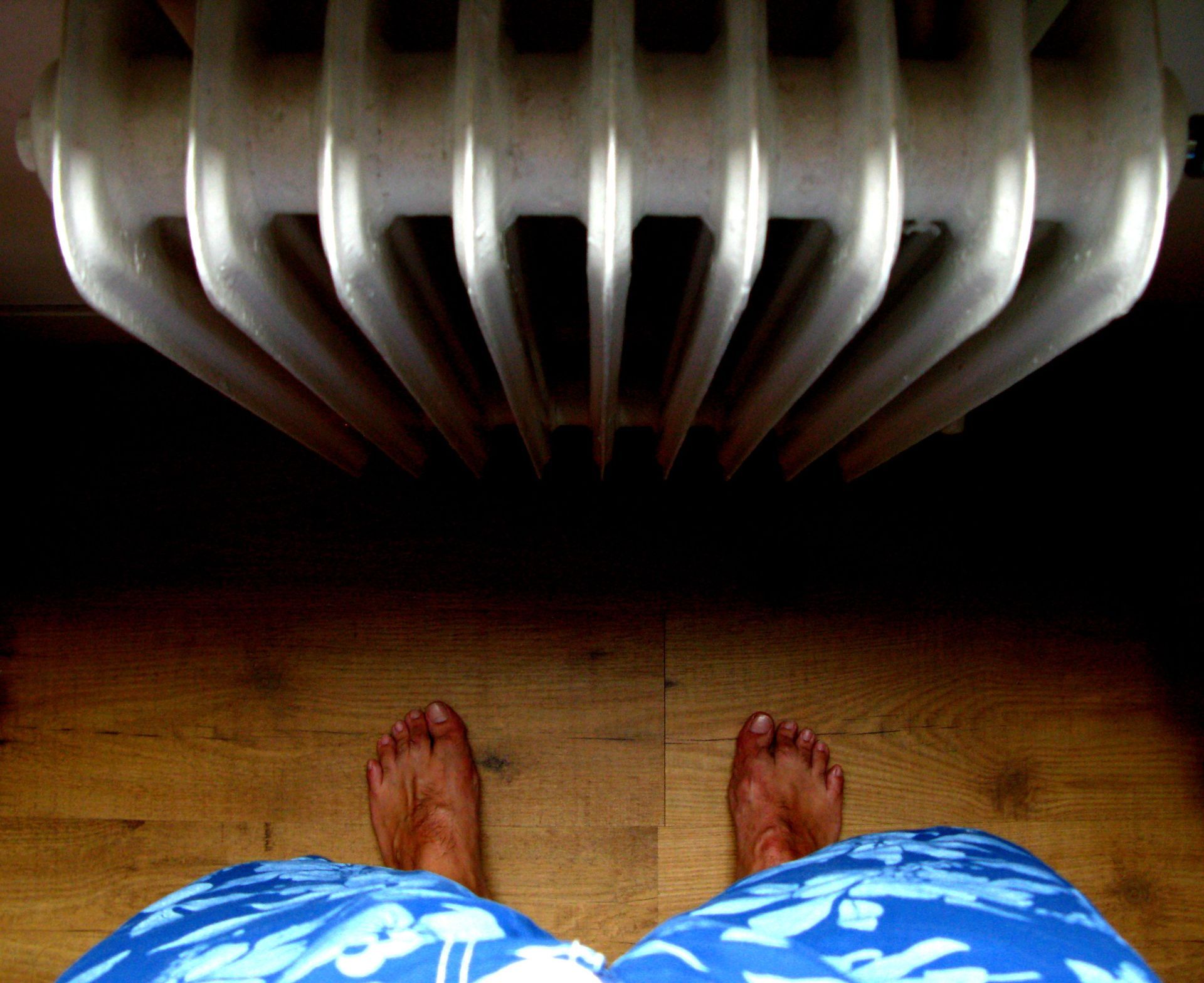 Bare feet in blue patterned shorts standing by a white radiator on a wooden floor