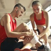 Two athletes in red sportswear checking a wrapped ankle on a gym floor