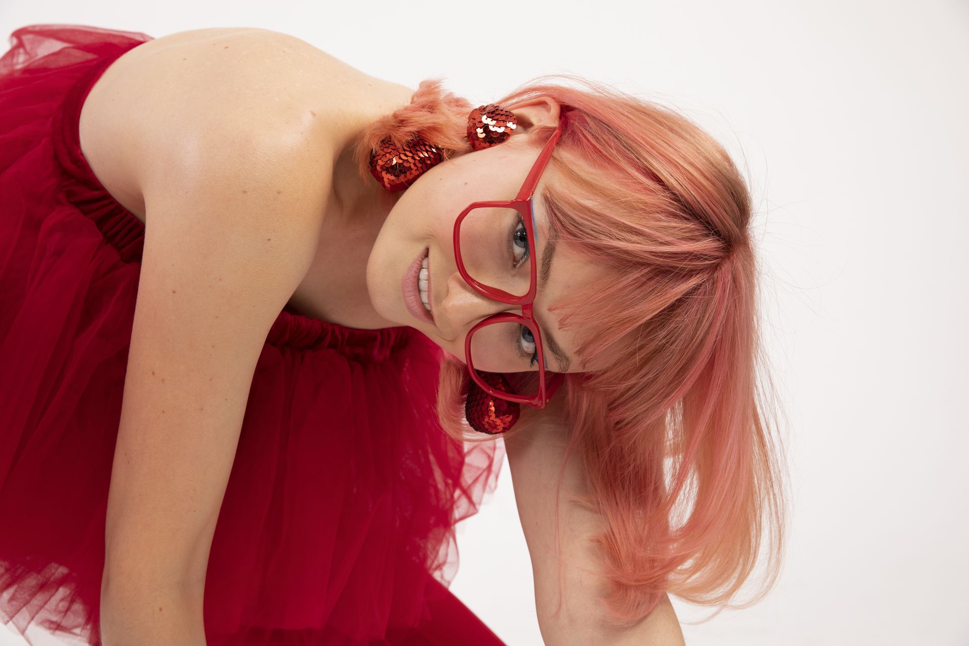 Una donna con tutù rosso, occhiali e orecchini si china, sorride e guarda verso la telecamera.