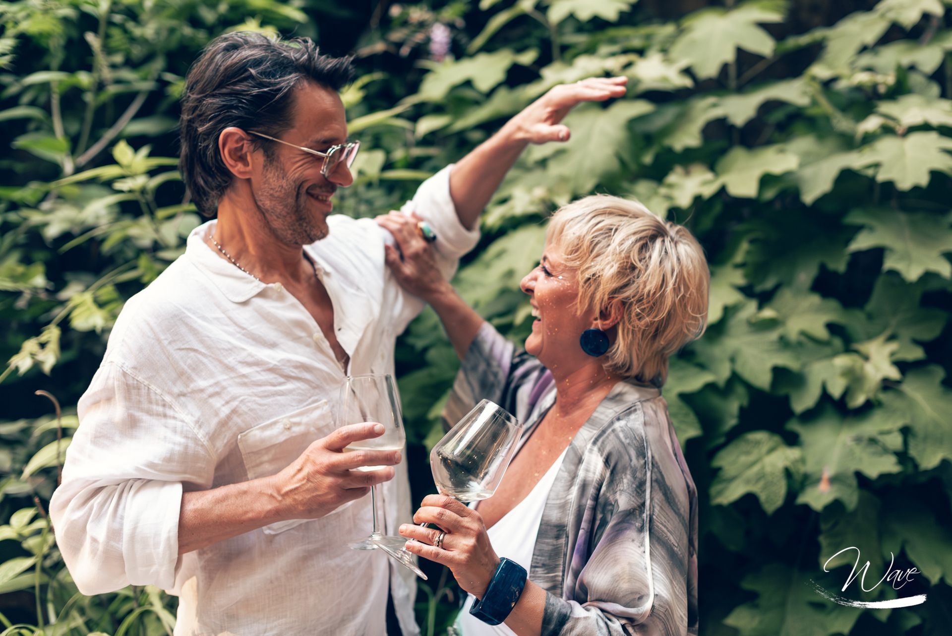 Coppia sorridente, abbracciata all'aperto, con in mano bicchieri di vino; l'uomo indica il verde.