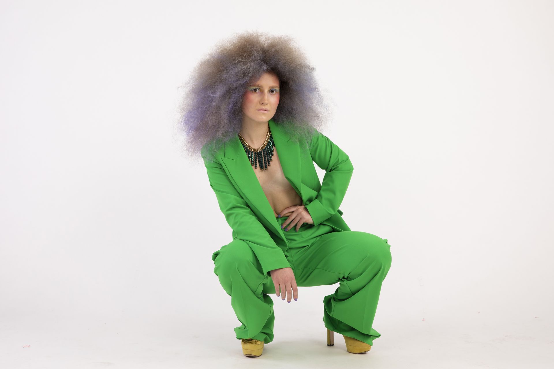 Persona con grande afro in abito verde, accovacciata, mano sul fianco. Sfondo bianco.