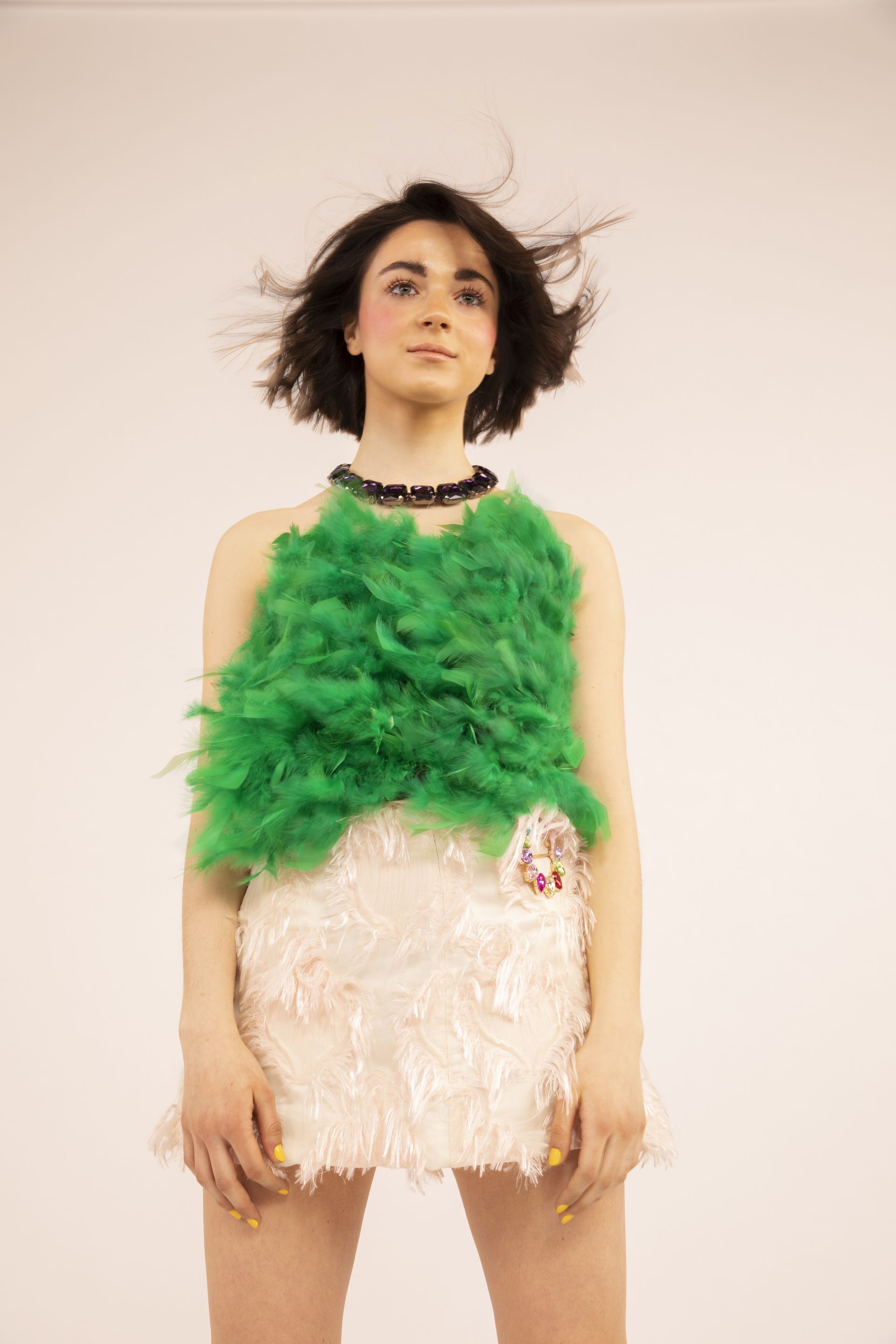 Donna con top di piume verdi e pantaloncini scintillanti, capelli al vento, su sfondo bianco.