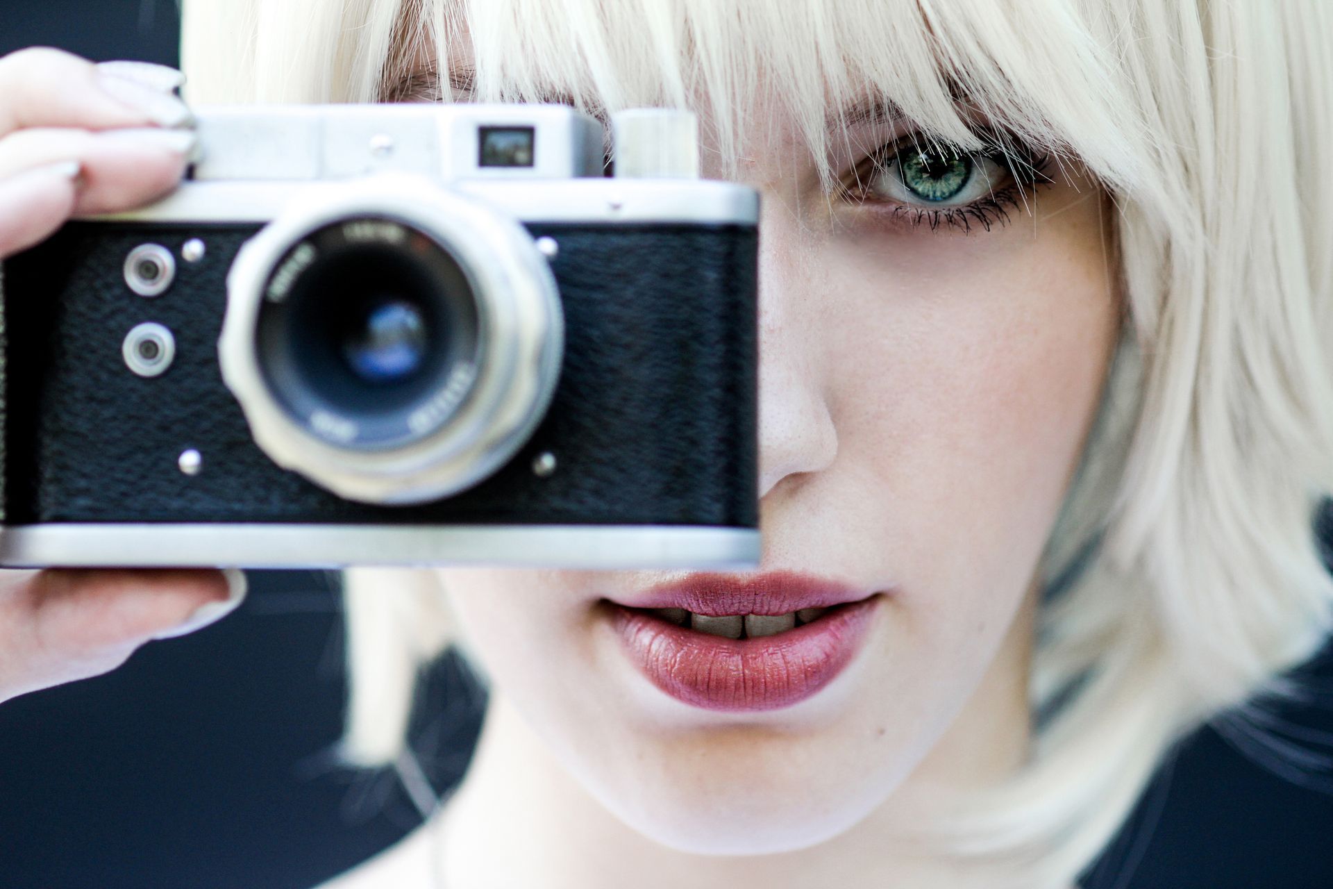 Woman with blonde hair holding a black and silver camera, looking through it with green eyes.