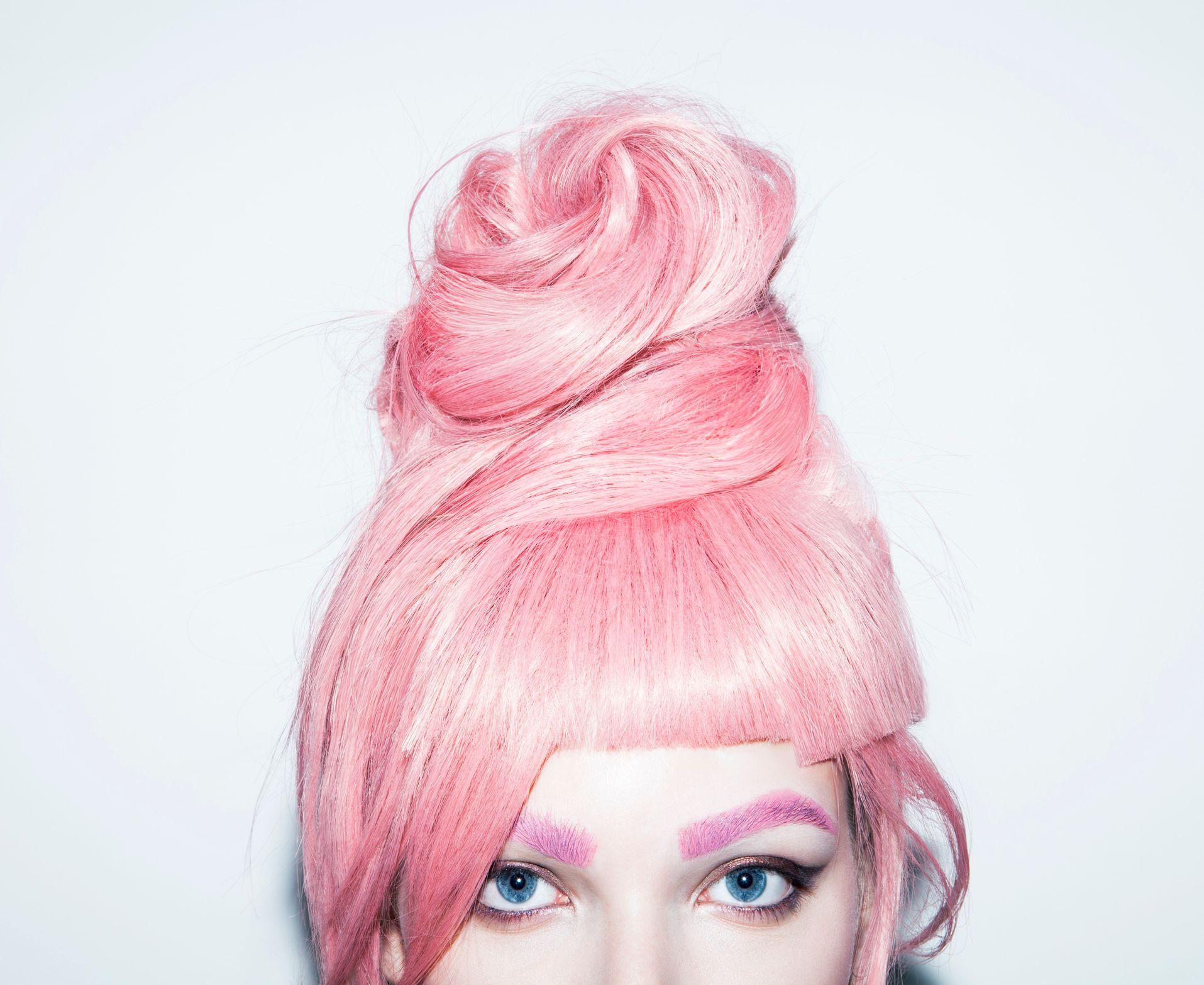 Woman with pink hair styled in a high bun and matching eyebrows, blue eyes, looking forward.