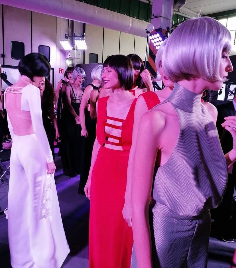 Modelle in abiti e caschetti nel backstage di una sfilata di moda, illuminate da luci intense.