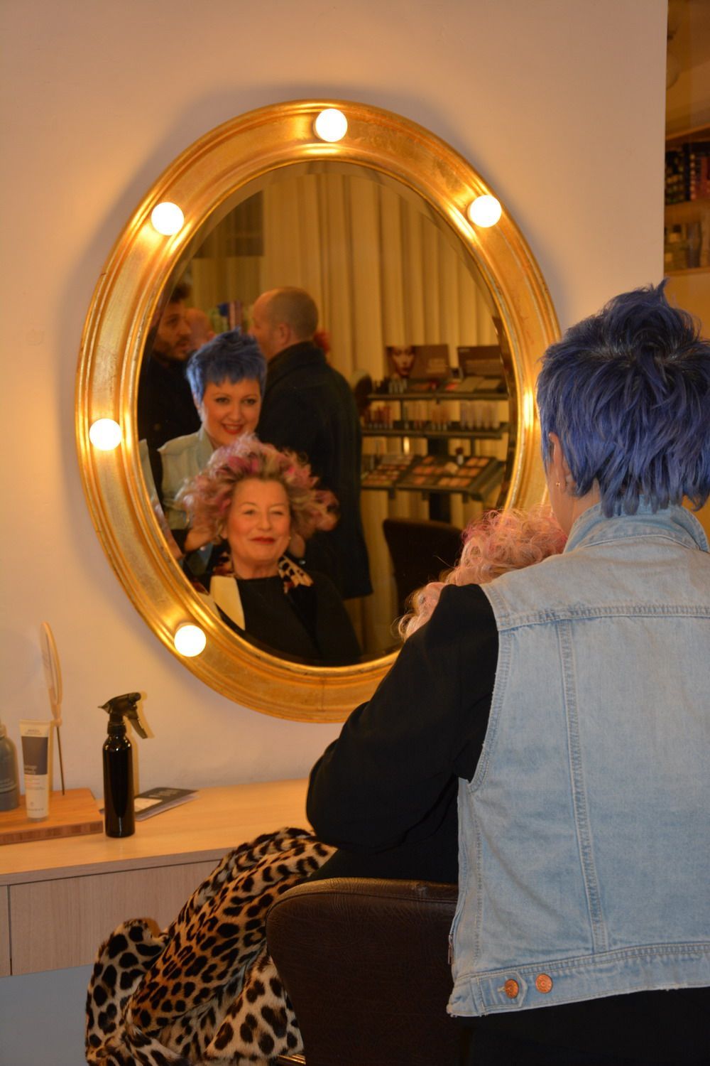 Una donna con i capelli blu in un salone di bellezza osserva il suo riflesso in uno specchio ovale illuminato;
