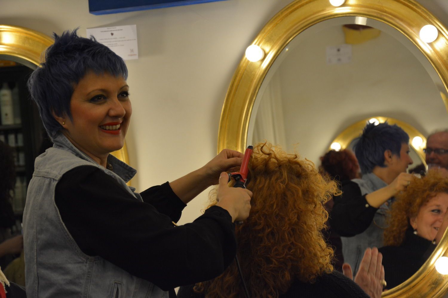 Parrucchiere con i capelli blu che arriccia i capelli rossi e ricci di una cliente davanti a uno specchio illuminato.