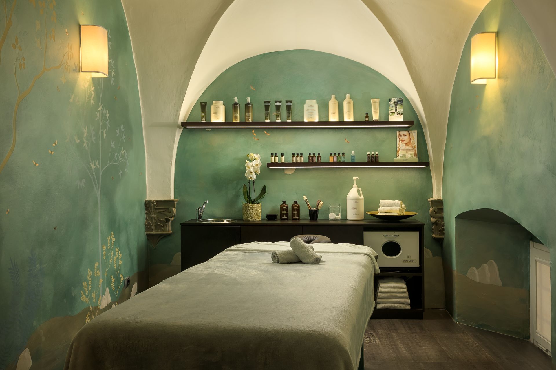 Sala benessere con lettino per massaggi, pareti color verde acqua, soffitto a volta e prodotti sugli scaffali.