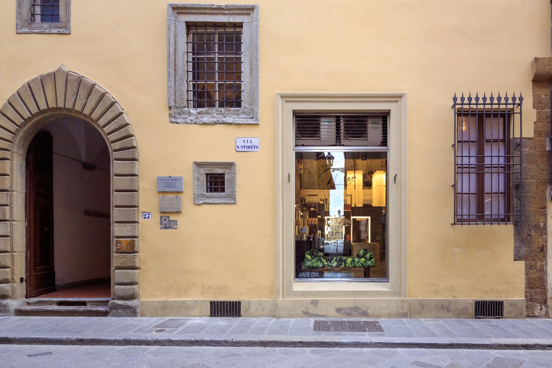Facciata gialla di un edificio con porta ad arco, vetrina che riflette la strada e finestre con sbarre.