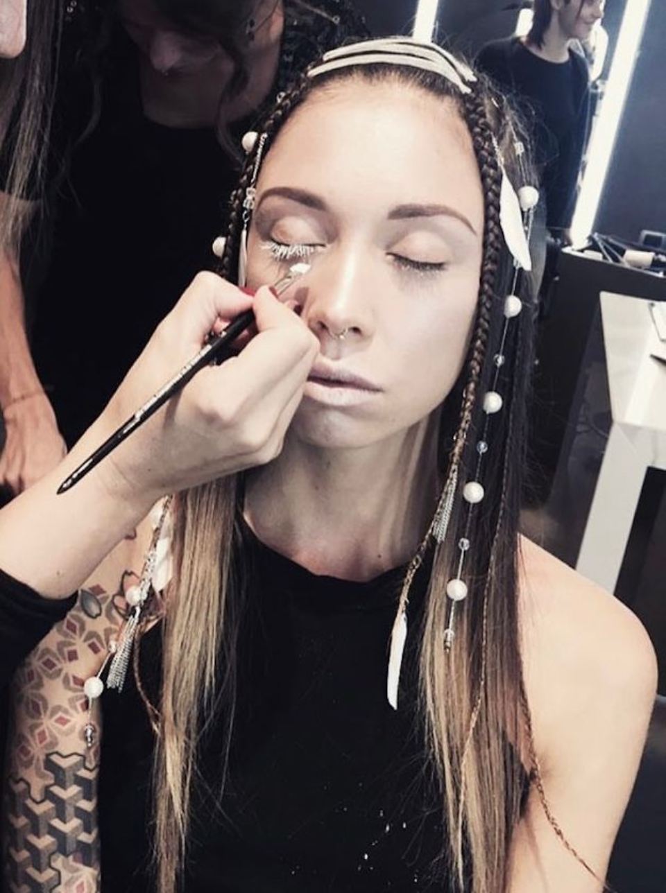 Una modella con il trucco applicato, capelli intrecciati con perle e piume. All'interno, una truccatrice.