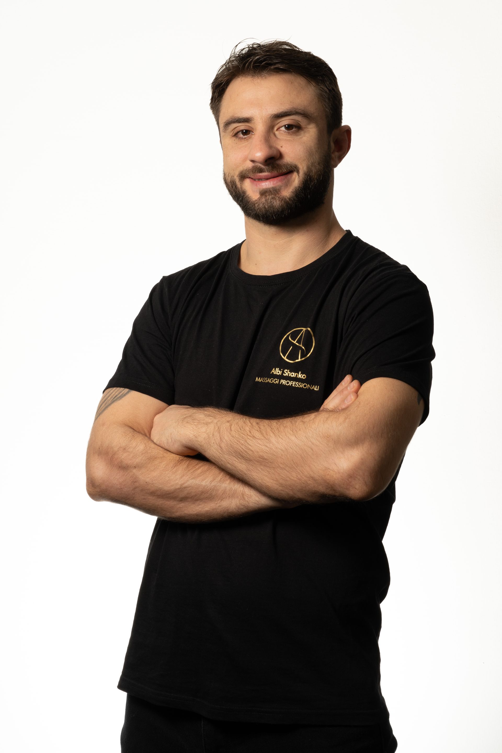 Uomo con le braccia incrociate, sorridente, che indossa una maglietta nera con logo dorato, su sfondo bianco.