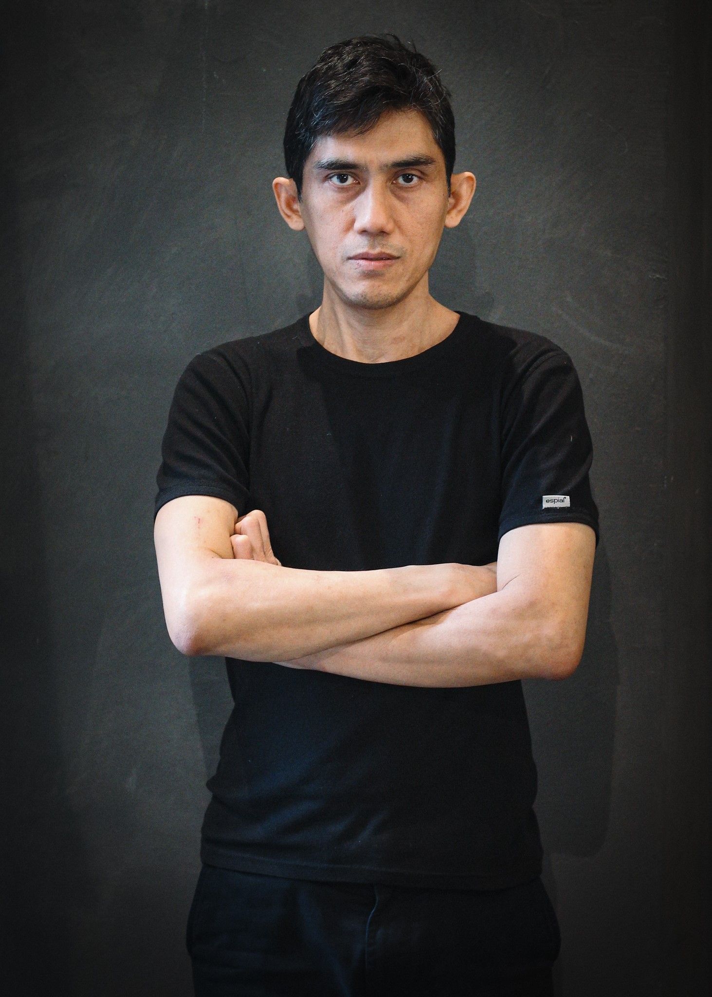 Uomo con le braccia incrociate, capelli scuri, maglietta nera, davanti a uno sfondo grigio scuro, che guarda la telecamera.