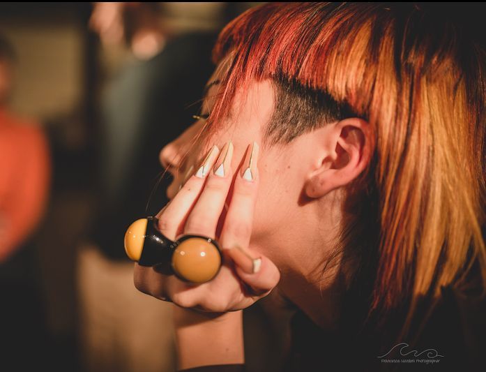 Woman with orange hair and shaved side, touching face with long nails and large ring.