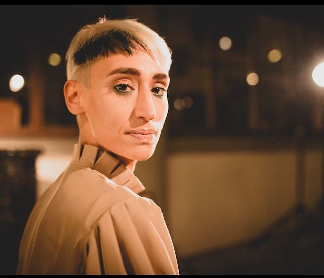 Person with short blonde hair, tan top, looking over their shoulder at night with bokeh lights.