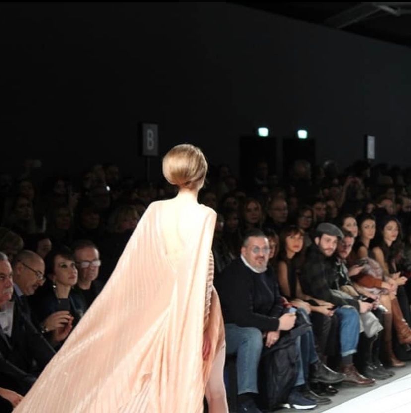 Modella in passerella con mantello rosa; il pubblico osserva dai posti a sedere.