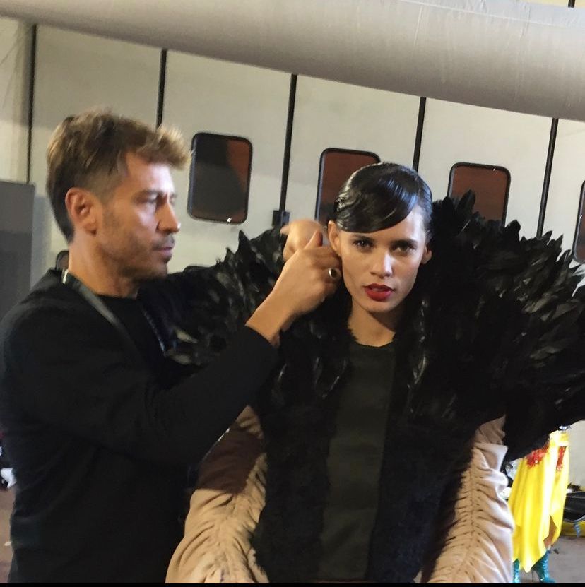 Man adjusting a feathered garment on a model backstage. Dark clothing, neutral tones.