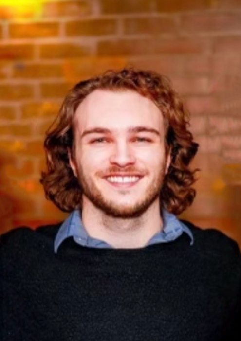 A man with long hair and a beard is smiling in front of a brick wall