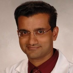 A man wearing glasses and a white coat is smiling for the camera.