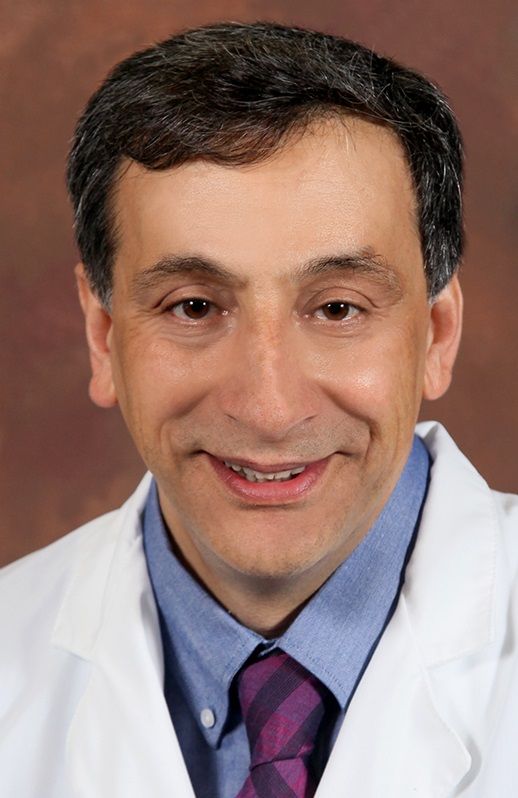 A man in a white coat and purple tie smiles for the camera