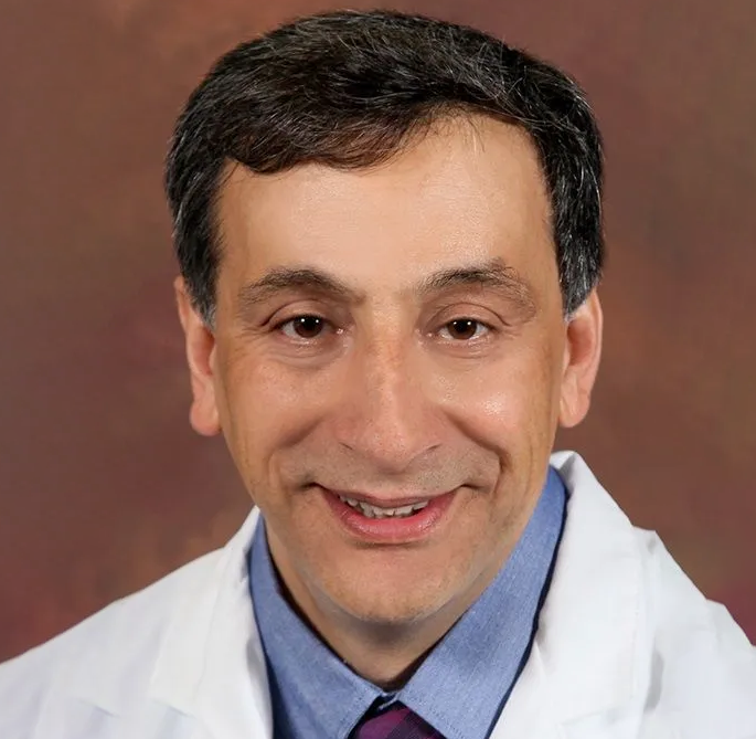 A man in a white coat and blue shirt is smiling for the camera.